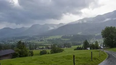 KI-Symbolbild: Dunkle Wolken ziehen über ein grünes Bergtal mit einer nassen Straße.