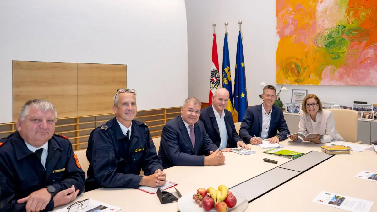 Menschen sitzen bei einem Arbeitsgespräch an einem langen Tisch, hinter ihnen stehen Fahnen.