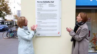 Zwei Frauen betrachten die Namenstafel am Herma-Nothelfer-Hof in Wien.