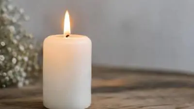 KI-Symbolbild: Eine weiße Kerze brennt auf einem Holztisch neben unscharfen Blumen.