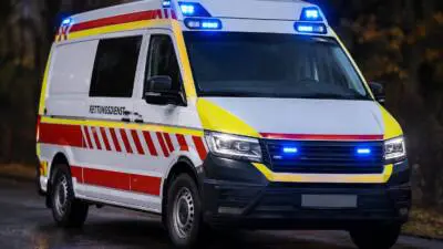 KI-generiertes Bild: Ein weißer Rettungsdienstwagen mit roten und gelben Markierungen und eingeschaltetem Blaulicht auf nasser Straße.