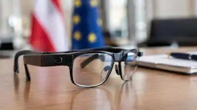 Schwarze Brille mit integrierter Kamera liegt auf einem Holztisch vor unscharfen Flaggen.