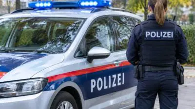 KI-generiertes Bild: Eine Polizistin steht neben einem weiß-blauen Polizeiauto mit eingeschaltetem Blaulicht.