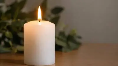 KI-Symbolbild: Eine brennende weiße Stumpenkerze steht auf einem Holztisch.