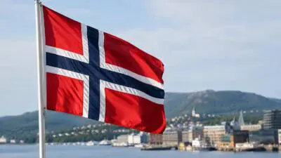 KI-Symbolbild: Eine norwegische Flagge weht vor einer Küstenstadt mit Bergen.