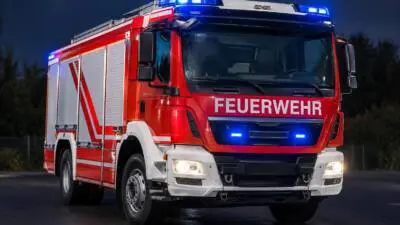 KI-generiertes Bild: Ein rotes Feuerwehrfahrzeug mit eingeschaltetem Blaulicht auf einer dunklen Straße.