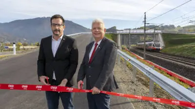 Zwei Männer halten ein rotes ÖBB-Infra-Band vor einer Straßenbrücke; daneben fährt ein Zug.