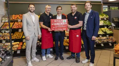 Männer halten ein rotes SPAR-Schild vor Obst- und Gemüseauslagen.