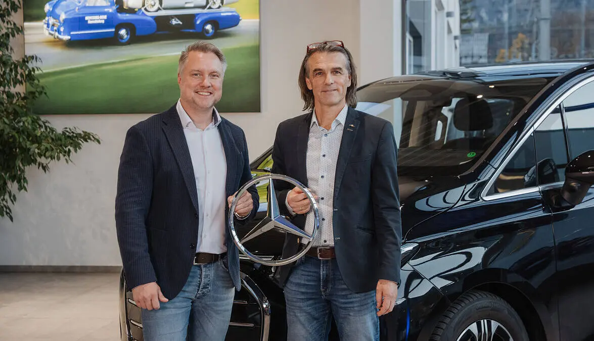 Zwei Männer halten einen Mercedes-Stern neben einem schwarzen Fahrzeug.