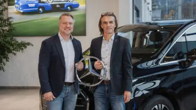 Zwei Männer halten einen Mercedes-Stern neben einem schwarzen Fahrzeug.