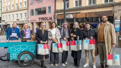 Menschen stehen mit bedruckten Stofftaschen neben einem türkisfarbenen Lastenrad; einige tragen auffälliges Gesichtsmake-up.