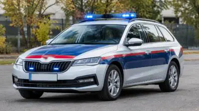 KI-generiertes Bild: Ein weißer Kombi mit blau-roter Lackierung und eingeschaltetem Blaulicht auf einer Straße.