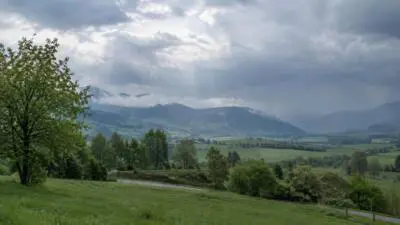KI-Symbolbild: Dunkle Wolken ziehen über ein grünes Tal mit bewaldeten Hügeln.