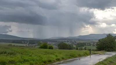 KI-Symbolbild: Regen fällt über eine grüne Hügellandschaft, eine nasse Straße liegt im Vordergrund.