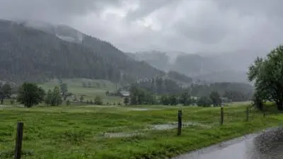 KI-Symbolbild: Eine regennasse Berglandschaft mit dunklen Wolken und Pfützen auf einer Wiese.