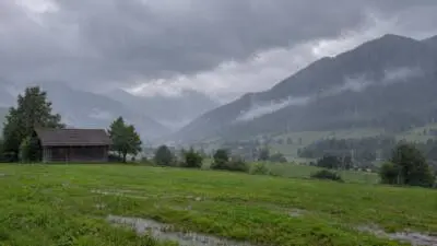 KI-Symbolbild: Regen fällt auf eine grüne Wiese vor einem bewölkten Bergtal.