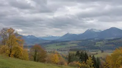 KI-generiertes Bild: Graue Wolken über einem herbstlichen Tal und bewaldeten Bergen.