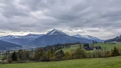 KI-Symbolbild: Bewölkte Berglandschaft mit grüner Wiese und schneebedeckten Gipfeln.