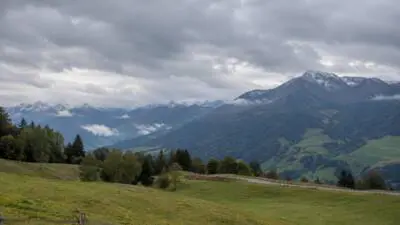 KI-Symbolbild: Dichte Wolken über schneebedeckten Berggipfeln und grünen Hängen.