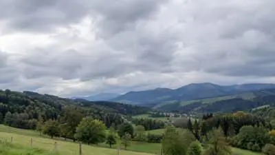 KI-Symbolbild: Graue Wolken über grünen Hügeln und bewaldeten Bergen.