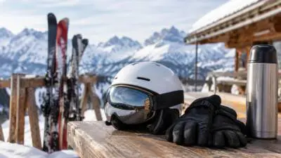 KI-generiertes Bild: Skihelm, Skibrille, Handschuhe und Thermoskanne auf einem Holztisch vor verschneiten Bergen.