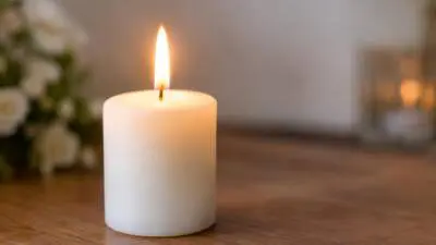 KI-generiertes Bild: Eine weiße brennende Kerze steht auf einem Holztisch neben unscharfen Blumen.