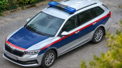 KI-Symbolbild: Ein silberner Kombi mit blau-roten Streifen und Blaulichtbalken steht auf einer Kiesfläche.