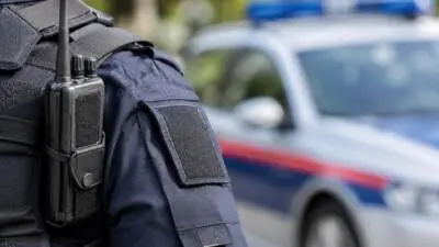 Polizeiuniformierter Oberkörper mit Funkgerät vor einem unscharfen Streifenwagen.