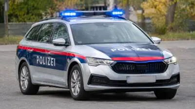 KI-generiertes Bild: Ein silberner Polizeikombi mit blau-roter Beschriftung und eingeschaltetem Blaulicht auf einer Straße.