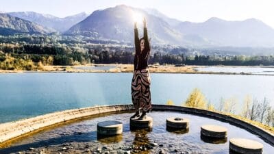 Frau mit ausgestreckten Armen zur Sonne steht auf Betonpfeiler in flachem Wasser vor See, Bergen und Herbstwald.