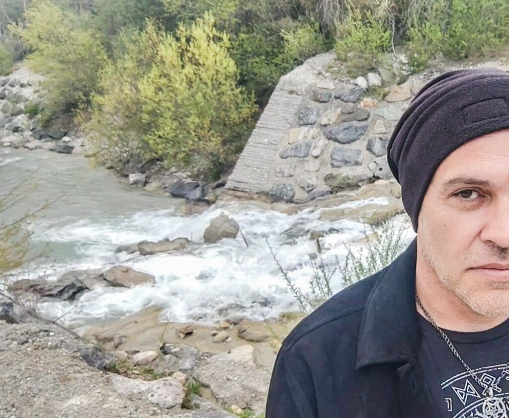 Mann mit dunkler Beanie und schwarzer Jacke steht vor rauschendem Fluss, Steindamm und grüner Vegetation.