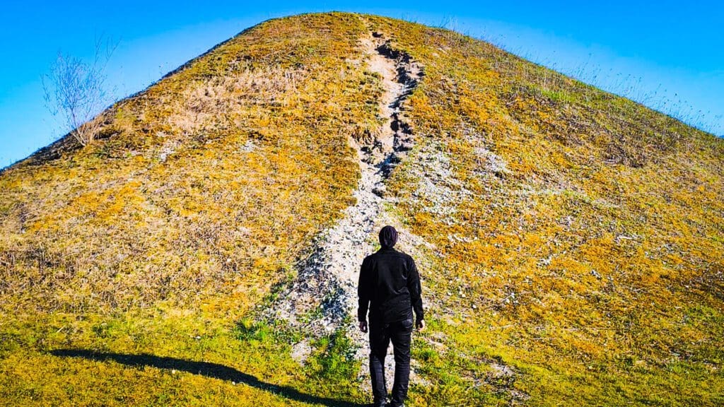 Mann in schwarzer Jacke und Hose steht mit dem Rücken zur Kamera auf grünem Hügel mit Pfad unter blauem Himmel.