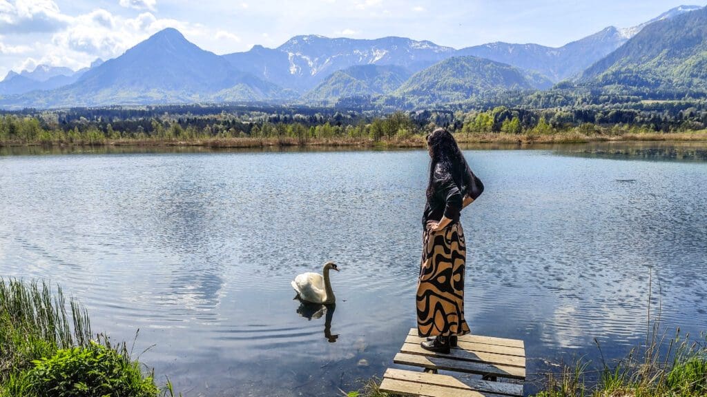 Frau mit langen schwarzen Haaren und buntem Rock steht auf Holzsteg am See und schaut zu weißem Schwan im Wasser mit Bergen im Hintergrund