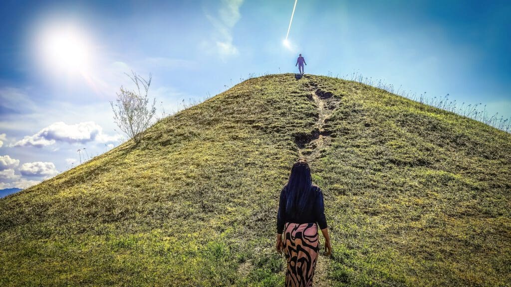 Eine Frau mit langem schwarzem Haar und gemustertem Rock geht einen Pfad eine grasbewachsene Anhöhe hinauf, vorne eine Gestalt oben bei strahlender Sonne.