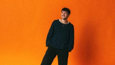 Ein lächelnder junger Mann mit dunklen Haaren und Bart vor orangem Hintergrund.