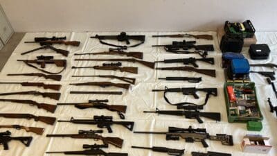 Viele Gewehre, Pistolen und Munitionsboxen auf weißer Plane in einem Raum neben grauen Schränken ausgelegt.