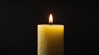 Eine brennende gelbe Kerze mit heller Flamme auf schwarzem Hintergrund als Symbol für Tod und Trauer.