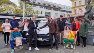 Gruppe von Personen mit After-Work-Markt-Plakaten posiert vor silbernem Auto und Statue am Benediktinerplatz.