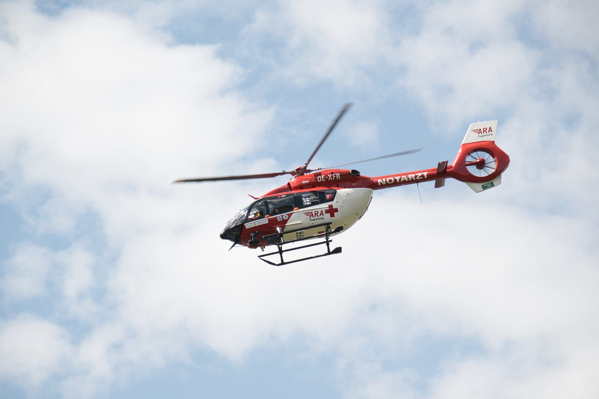 Roter und weißer Rettungshubschrauber der ARA Flugrettung fliegt vor blauem Himmel mit Wolken.