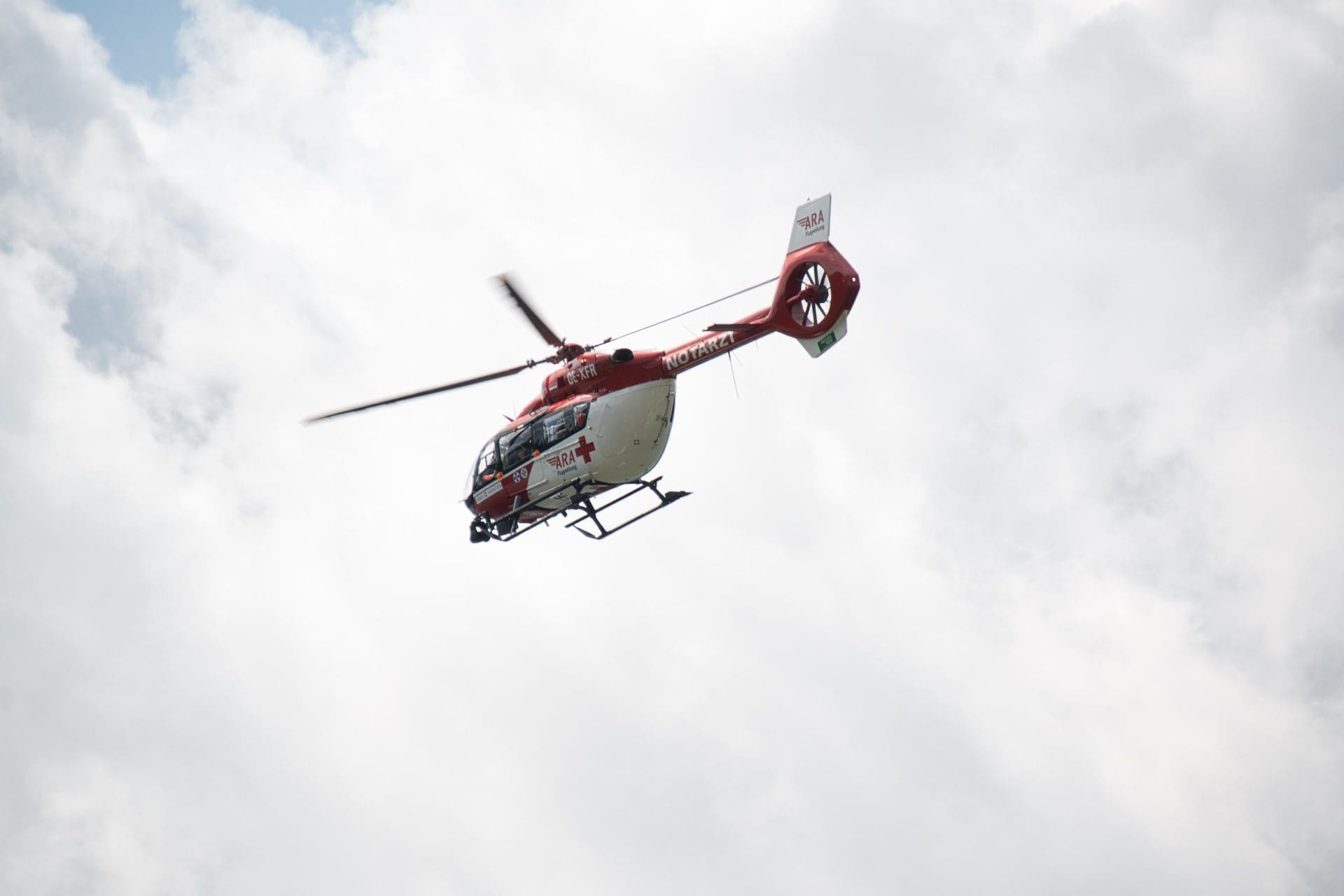 Ein roter und weißer Rettungshubschrauber der ARA Flugrettung fliegt vor einem bewölkten Himmel.