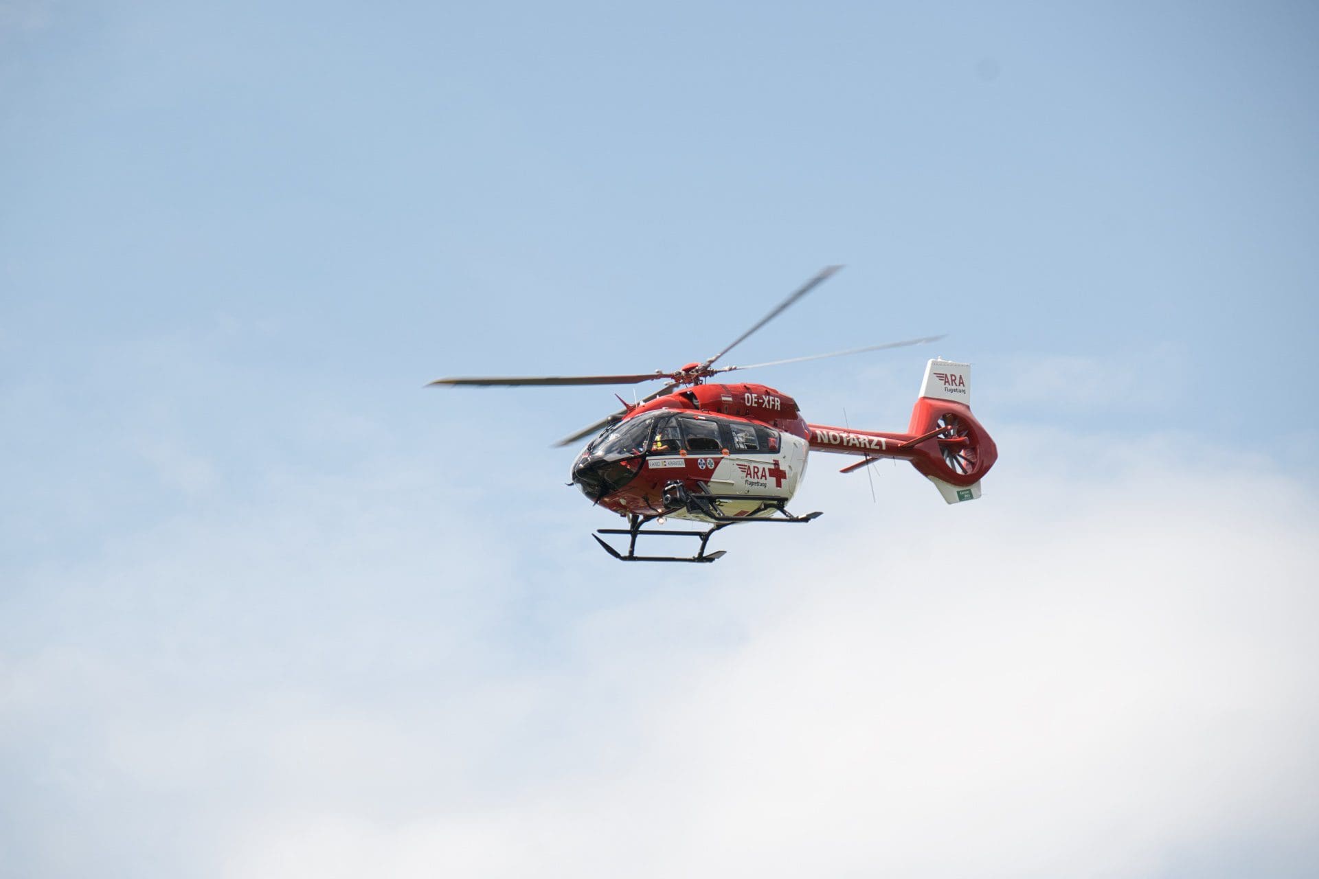 Roter und weißer Rettungshubschrauber der ARA Flugrettung mit NOTARZT-Beschriftung im Flug vor blauem Himmel