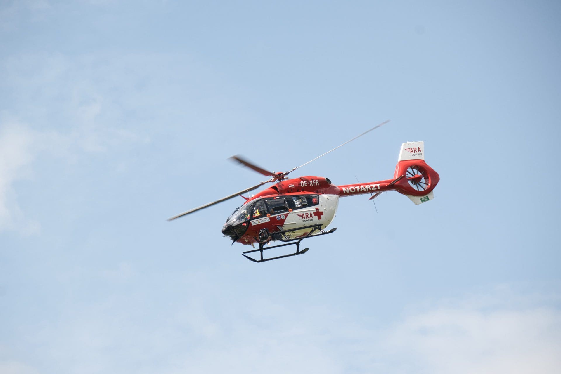 Roter und weißer Rettungshubschrauber der ARA Flugrettung mit NOTARZT-Beschriftung fliegt vor blauem Himmel