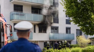 Feuerwehrleute und Polizisten vor einem Mehrparteienhaus mit Rauch aus einem Balkon