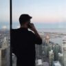 Ein Mann in schwarzem Hemd steht am Fenster und blickt auf die Skyline einer Stadt am Wasser.