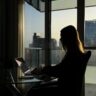 Silhouette einer Frau am Laptop vor großen Fenstern mit Stadtblick