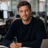Junger Mann mit braunen Locken und blauem Pullover sitzt am Schreibtisch mit Stift und Notizbuch