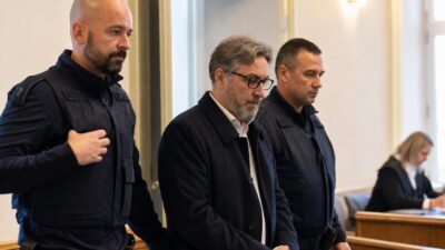 Ein grauhaariger Mann mit Brille steht zwischen zwei uniformierten Beamten in einem Gerichtssaal.