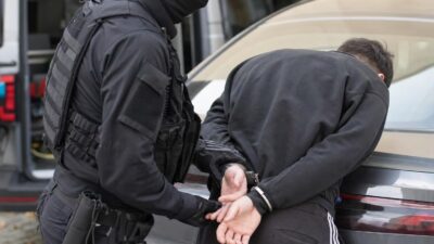 Cobra-Beamter in schwarzer Schutzausrüstung legt einem Verdächtigen Handschellen an einem Auto an