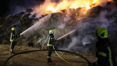 Feuerwehrleute in Schutzkleidung löschen mit Wasserschläuchen einen brennenden Strohballenstapel bei Nacht