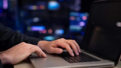 Hände einer Person tippen auf der Tastatur eines silbernen Laptops vor unscharfen Bildschirmen.